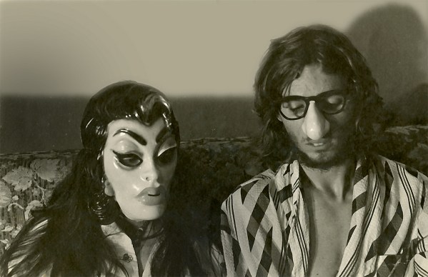 A couple sitting in bed, somewhat "John & Yoko" like, but wearing masks on their faces.